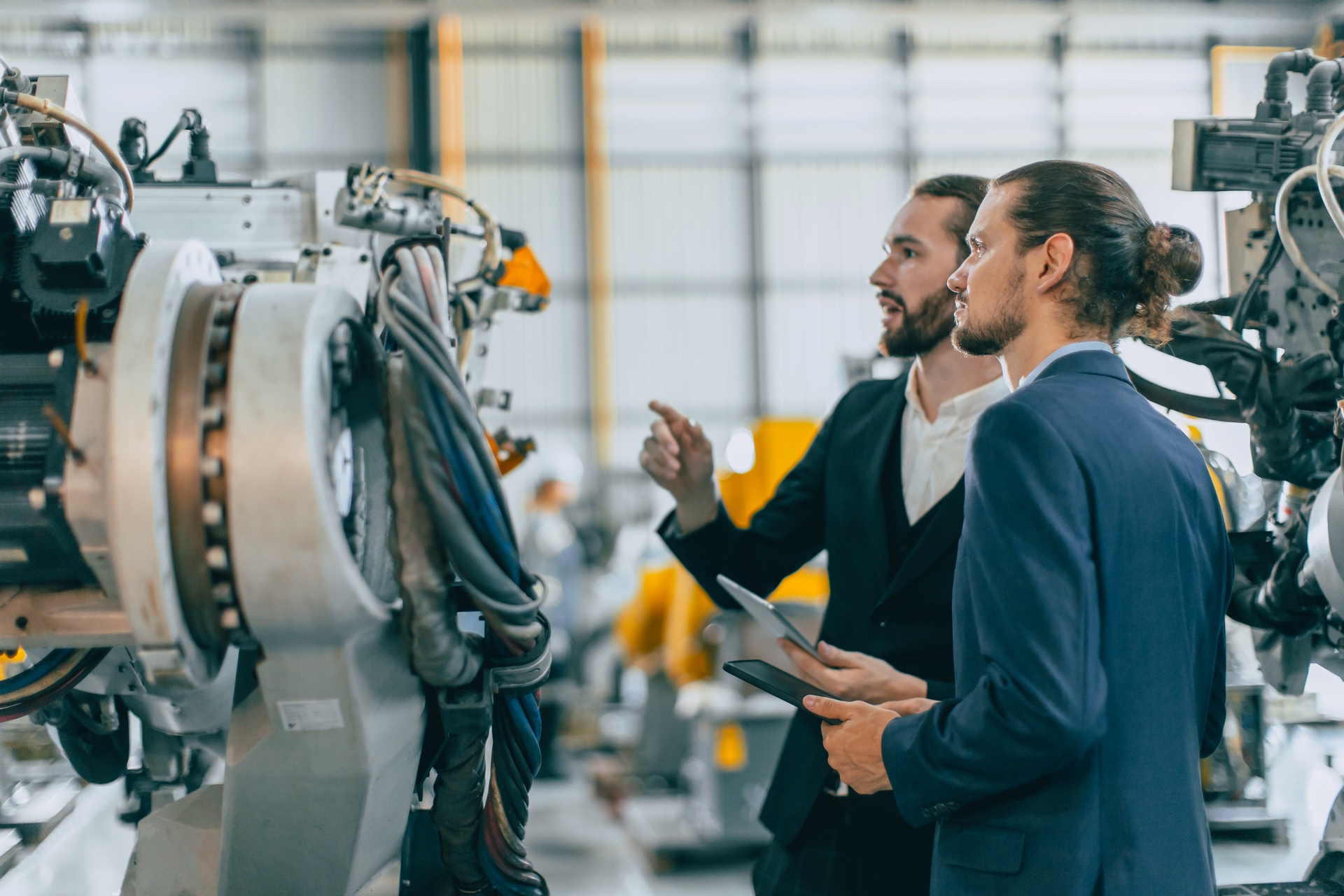 Two men in suits looking at a mechanical device in an industrial environment and discussing it.