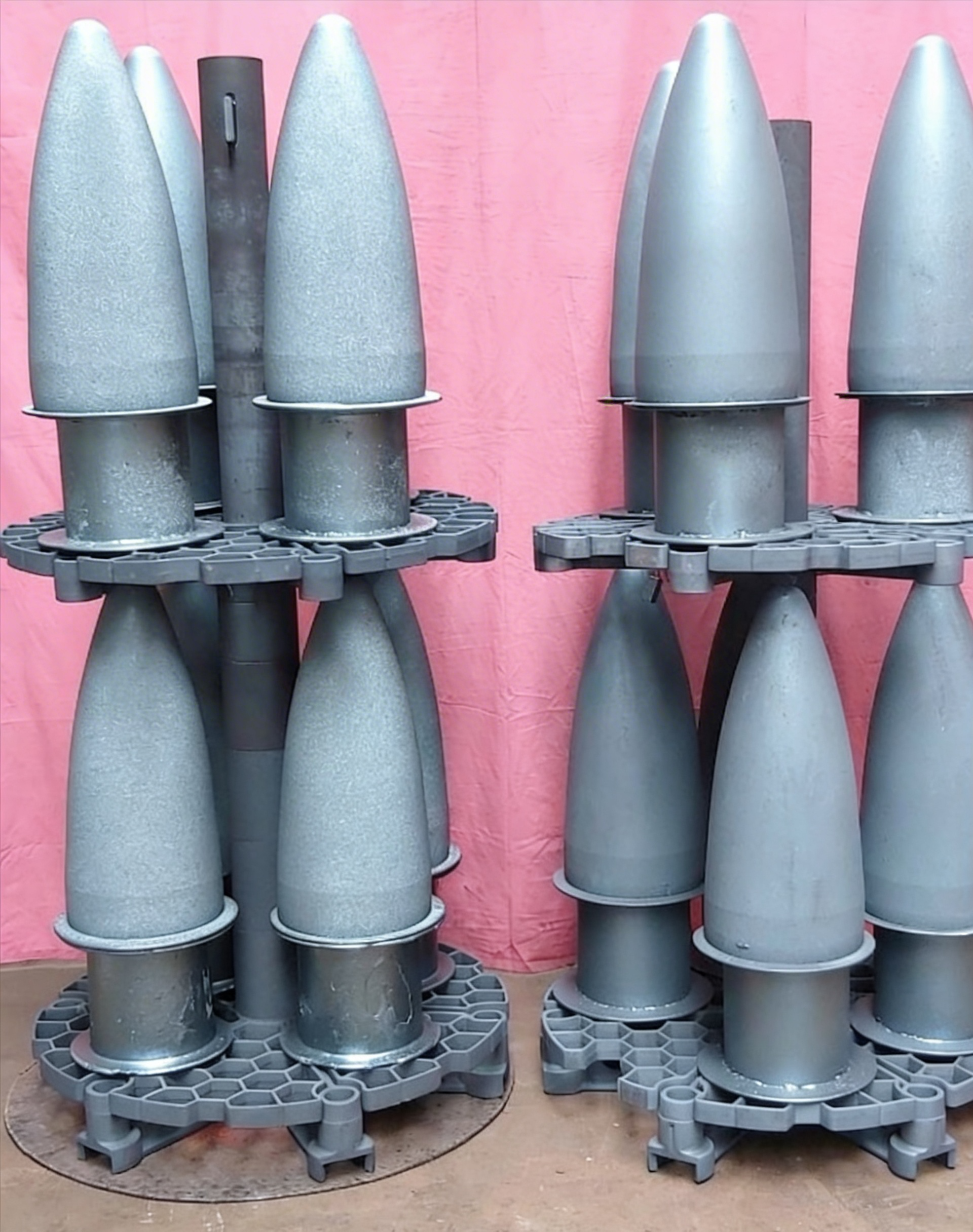 Zwei Ständer mit grauen, zylindrischen Objekten, die möglicherweise Munitionsgeschosse darstellen, vor rosa Hintergrund.