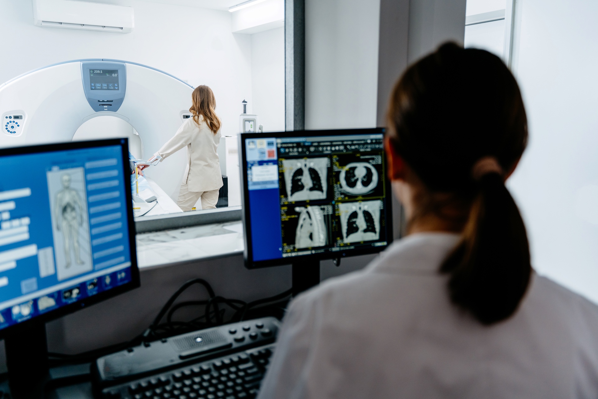 One person is standing in front of a CT scanner while a second person analyses X-ray images on the computer.